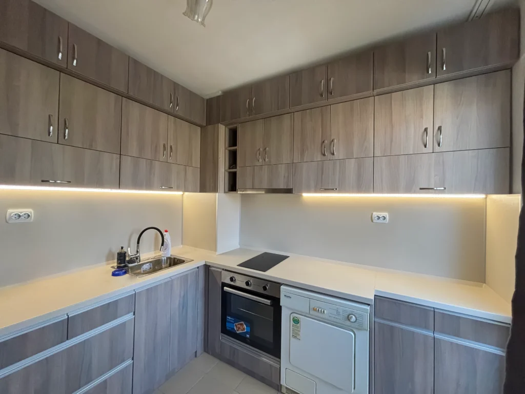 Пълен изглед на кухнята – осветена композиция в дървесни тонове и светъл плот Шампейн.